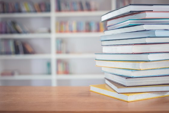 Im Vordergrund ein Stapel Bücher auf einem Schreibtisch, im Hintergrund ein Bücherregal